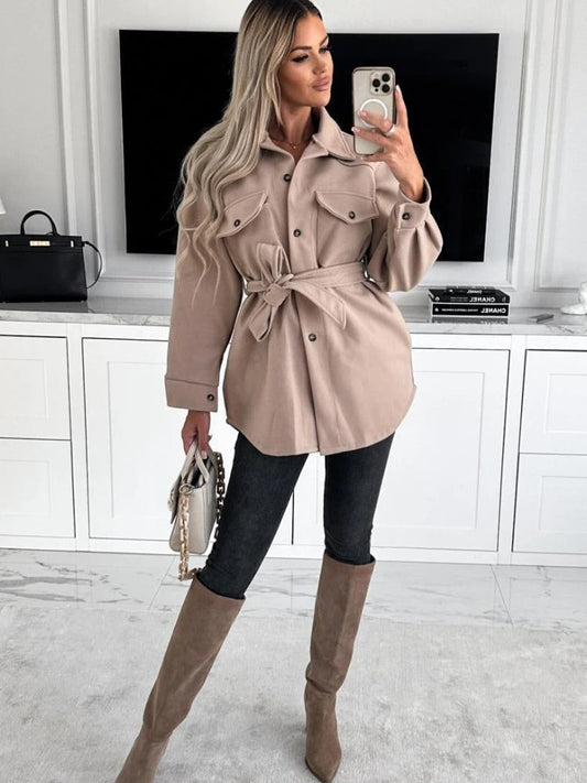 Frau in beiger Jacke mit Gürtel, schwarze Jeans, braune Stiefel, Selfie, modernes Wohnzimmer.