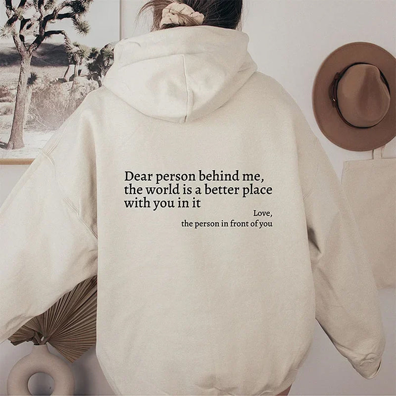 Frau in beigem Hoodie mit Aufschrift, Wanddekoration, Hut, Pflanzen im Hintergrund.