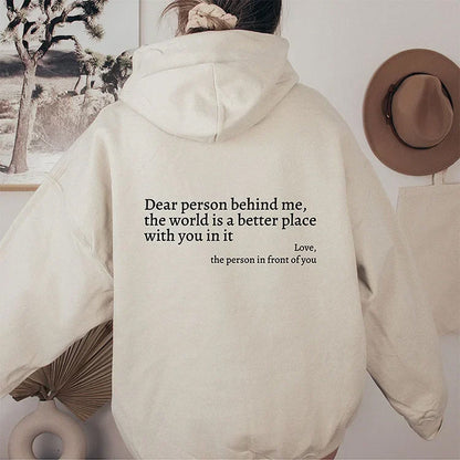 Frau in beigem Hoodie mit Aufschrift, Wanddekoration, Hut, Pflanzen im Hintergrund.
