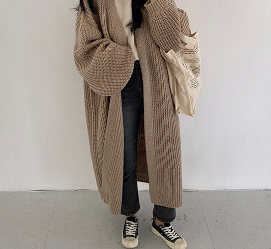 Frau in langem, beige Strickmantel, schwarze Jeans, Sneakers, hält Stofftasche.