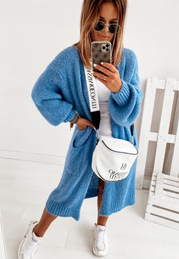 Frau in blauem Strickcardigan, weißem Top, Sonnenbrille, mit weißer Umhängetasche und Sneakers.