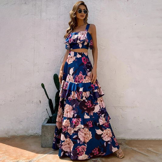 Frau in blauem, geblümtem Maxikleid mit Rüschen, Sonnenbrille, Sandalen, vor weißer Wand.