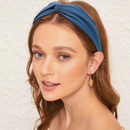 Frau mit blauem Stirnband, goldenen Ohrringen, braunen Haaren, lächelnd vor weißem Hintergrund.