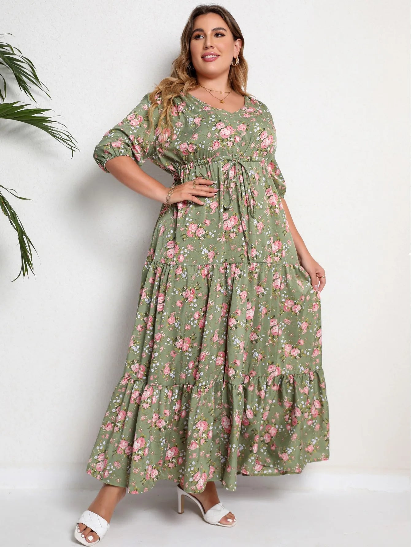 Frau im grünen, geblümten Maxikleid, Plus-Size, mit weißen High Heels, lächelt.