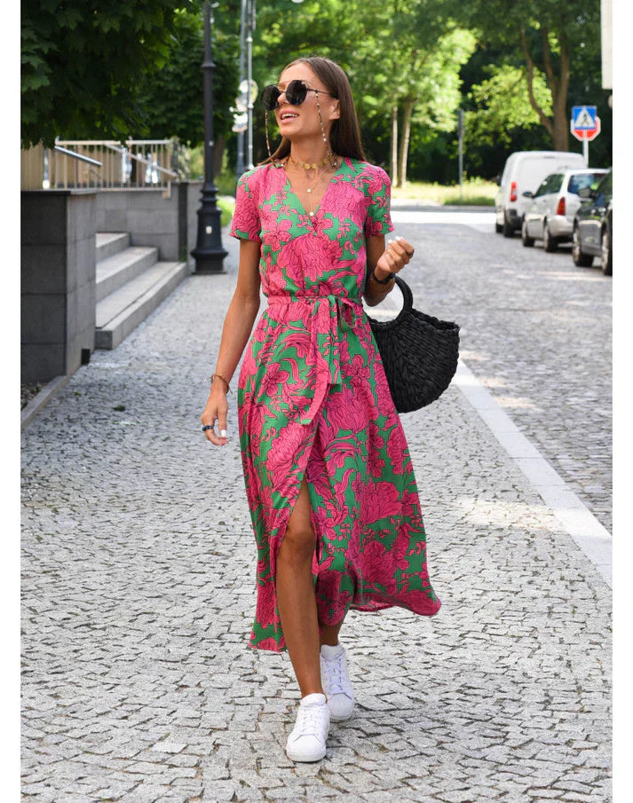Frau in pink-grünem Sommerkleid mit Korbtasche und weißen Sneakers auf gepflasterter Straße.