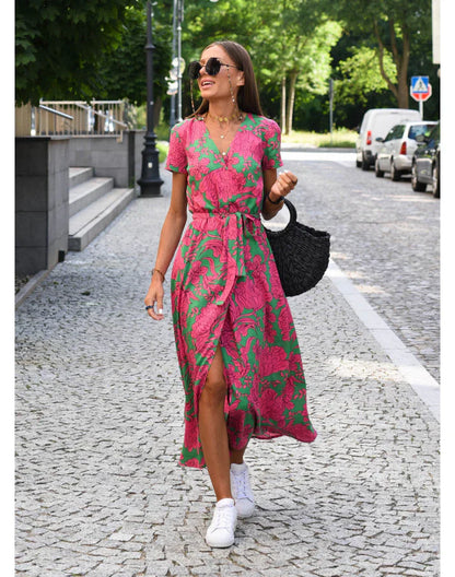 Frau in pink-grünem Sommerkleid mit Korbtasche und weißen Sneakers auf gepflasterter Straße.