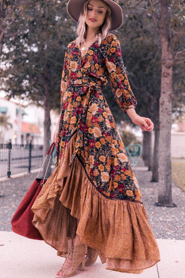 Frau in langem, geblümtem Kleid mit Hut und roter Tasche im Freien.