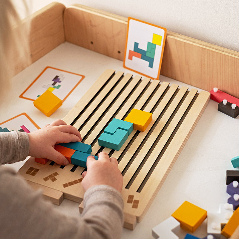 Kinderhände schieben bunte Holzsteine in die Schlitze des Bretts, im Hintergrund stehen Vorlagenkarten mit Mustern. 3