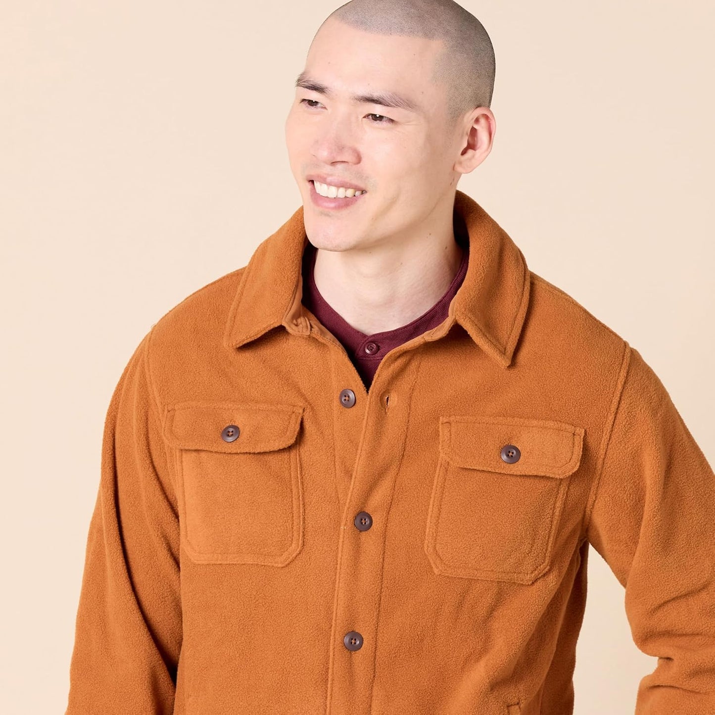 Mann in brauner Fleecejacke mit Knöpfen, Hemdkragen und Brusttaschen, lächelnd vor beigem Hintergrund.