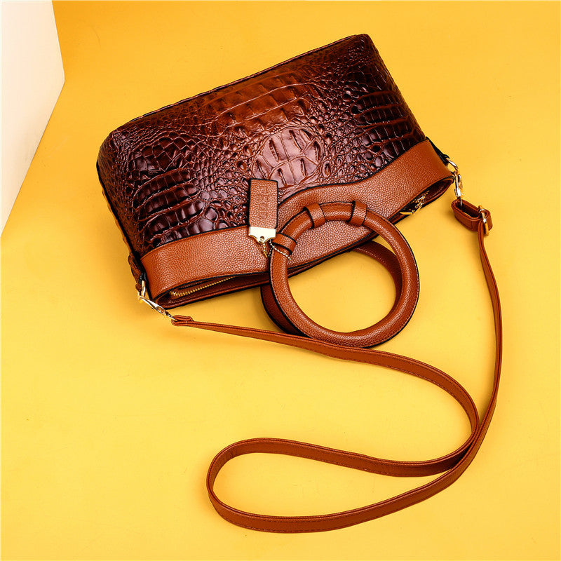Braune Krokodilleder-Handtasche mit Rundgriff und Schulterriemen auf gelbem Hintergrund.
