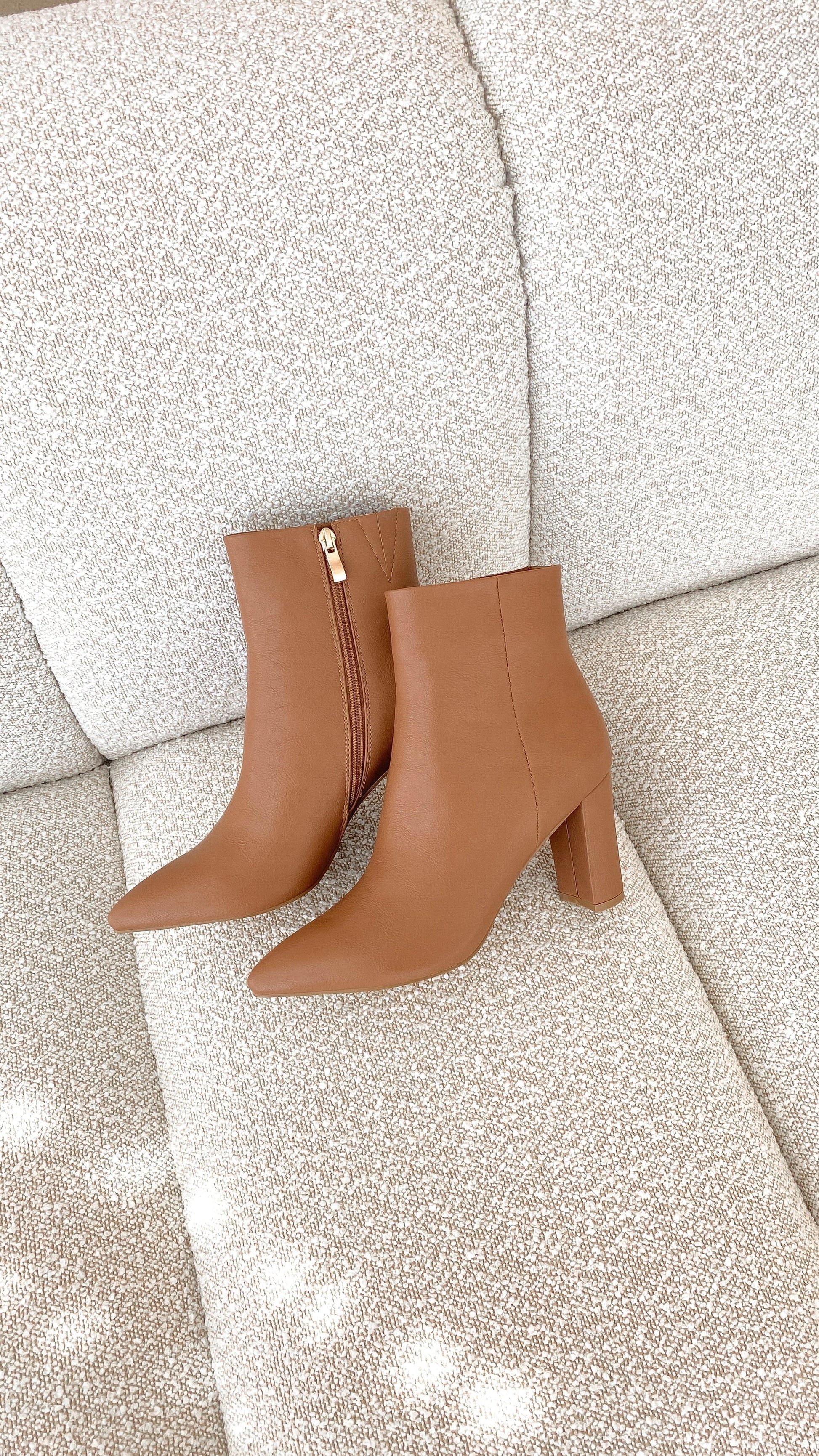 Braune Damenstiefeletten mit Absatz, Reißverschluss, Lederoptik, auf beigem Sofa.