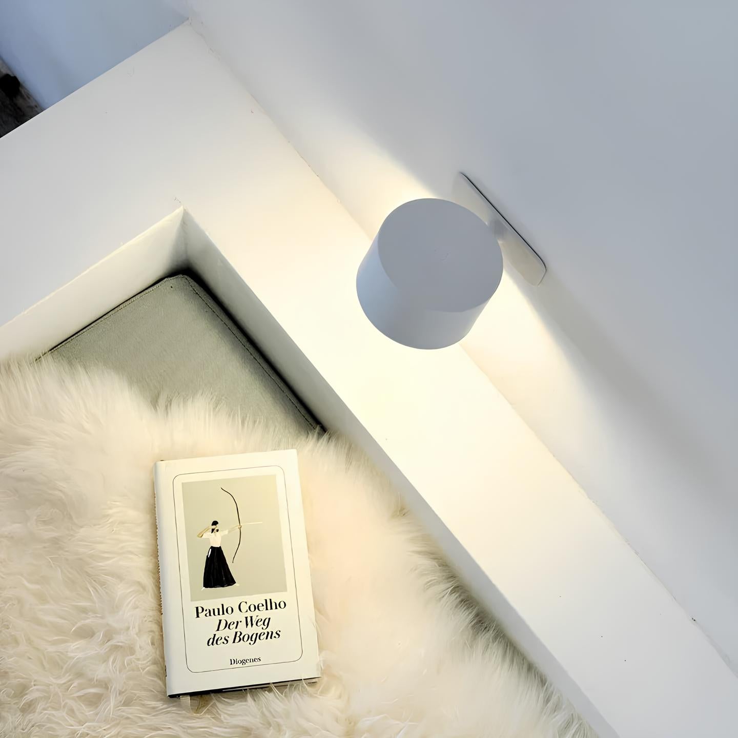 Buch "Der Weg des Bogens" von Paulo Coelho auf weißem Fell neben moderner Wandlampe.