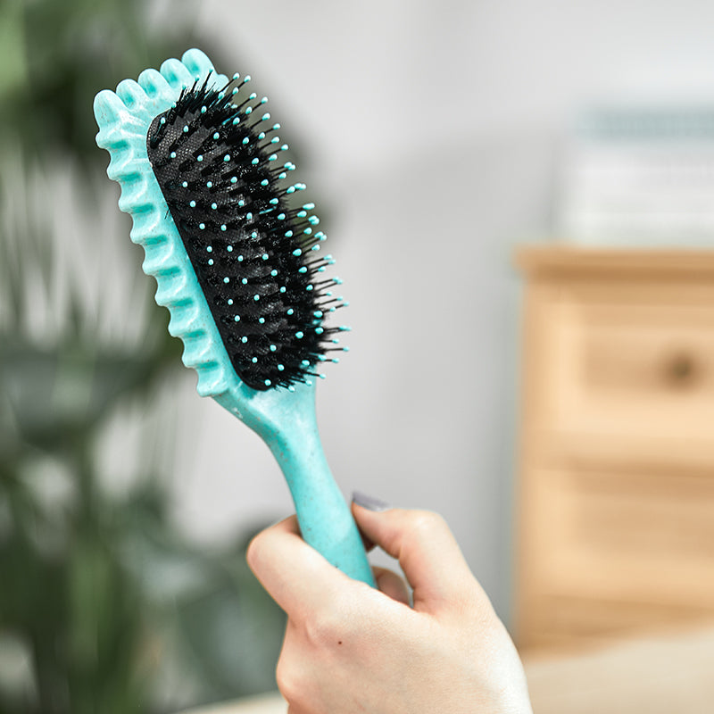 Türkise Haarbürste mit schwarzen Borsten, ergonomisch, in Hand gehalten, vor unscharfem Hintergrund.