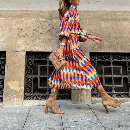 Frau in bunter Midi-Kleid mit geometrischem Muster, braune Stiefel, beige Tasche, Stadtstraße.