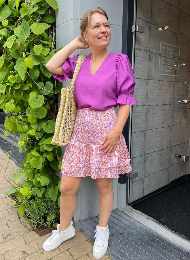Frau in lila Bluse und bunter Rock, trägt Strohtasche, weiße Sneaker, steht vor grünem Hintergrund.