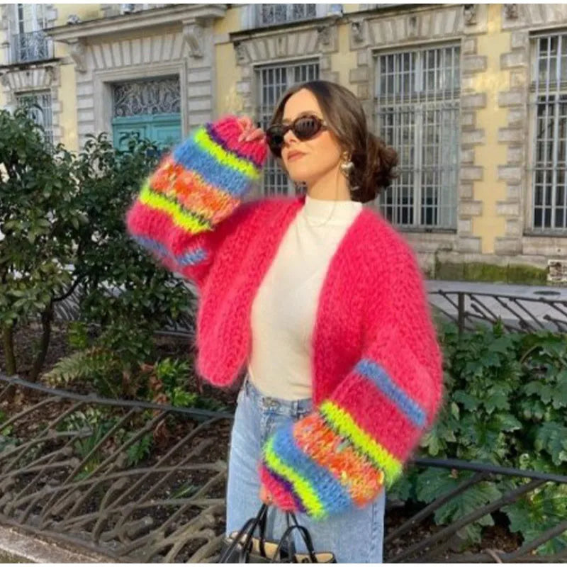 Frau in pinker Strickjacke mit bunten Streifen, Sonnenbrille, draußen vor historischem Gebäude.