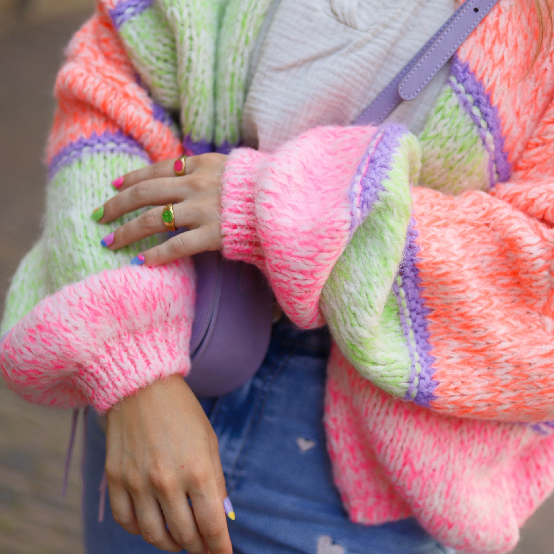 Frau in bunter Strickjacke, lila Tasche, Jeansrock, bunte Ringe und Nägel.