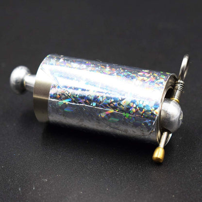 Nahaufnahme der aufgerollten Stabrolle mit holografisch schimmernder Folie, Metallknauf, Feder, Ring und goldenem Stift auf 5