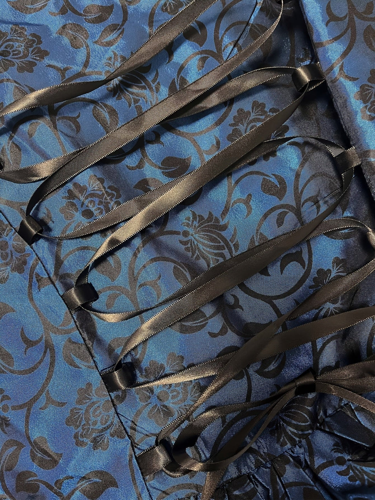 Detailaufnahme der schwarzen Satinband-Schnürung auf dem blauen Stoff mit schwarzem Blumenranken-Muster. 5