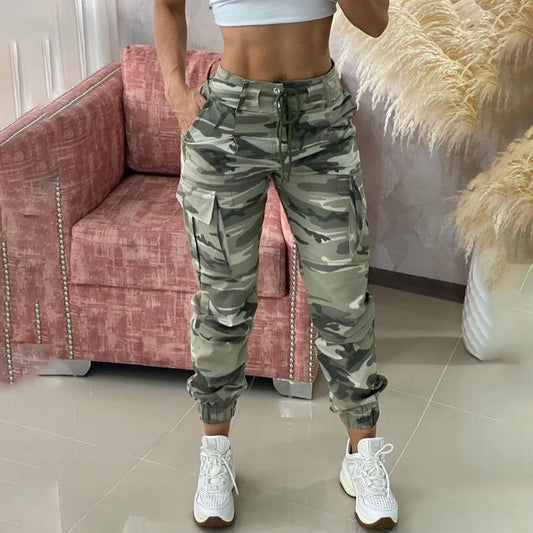 Frau in Camouflage-Cargohose und weißen Sneakers vor rosa Sofa.
