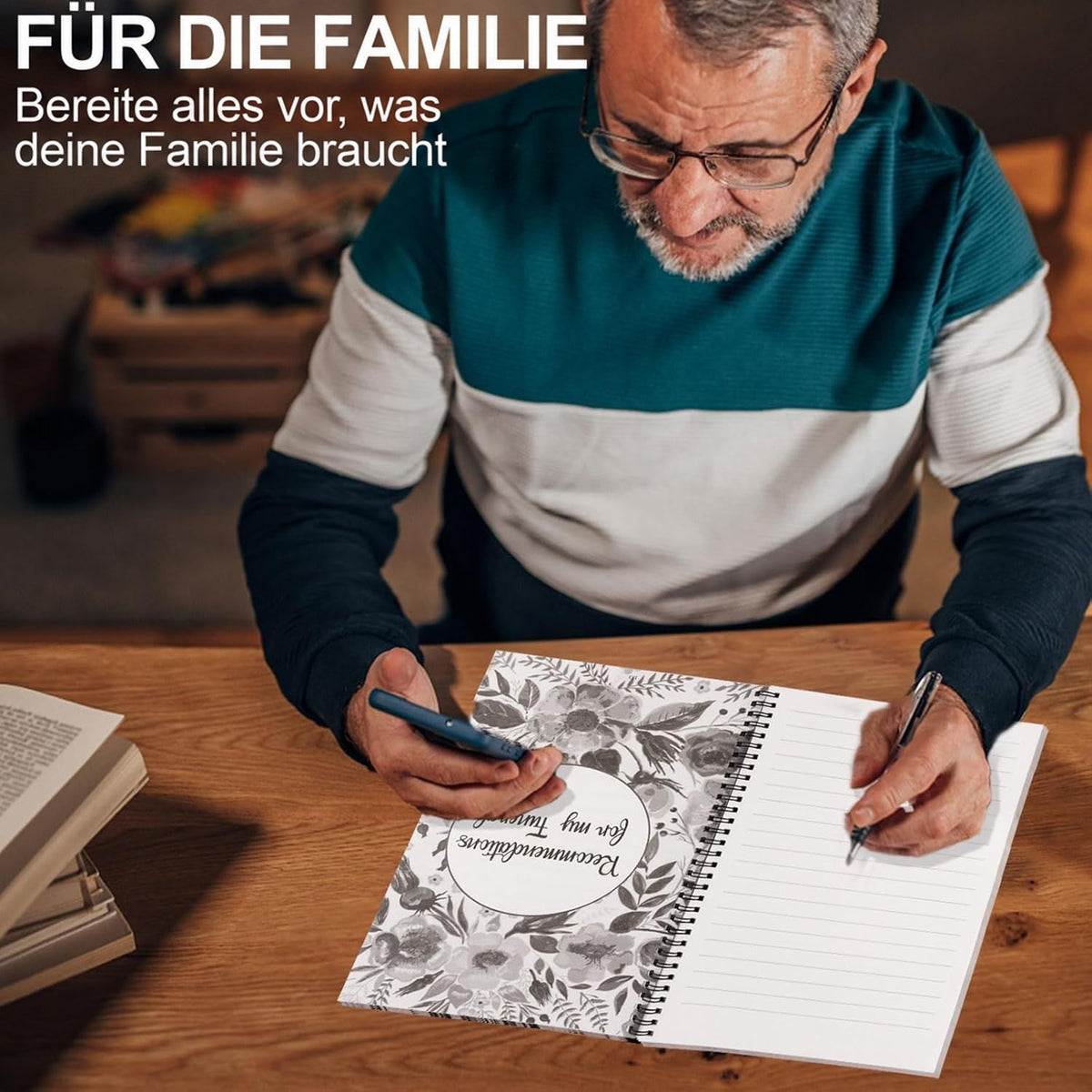 Älterer Mann mit Brille sitzt am Holztisch, hält ein Smartphone und schreibt mit Stift in den aufgeschlagenen Planer mit Blumenseite. 4