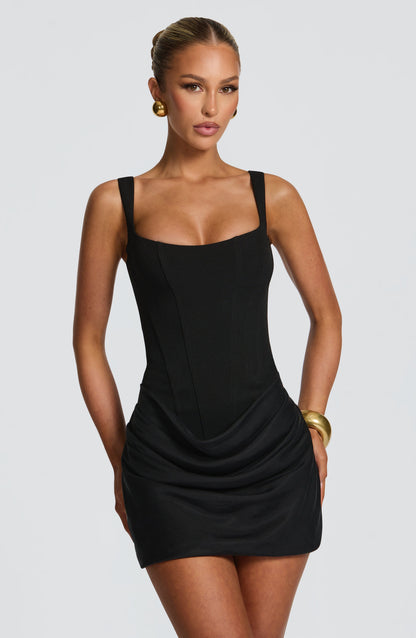 Schwarzes Minikleid frontal im Brustbild, mit quadratischem Ausschnitt, Korsagennähten und drapiertem Rockteil, dazu goldene Ohrringe. 10