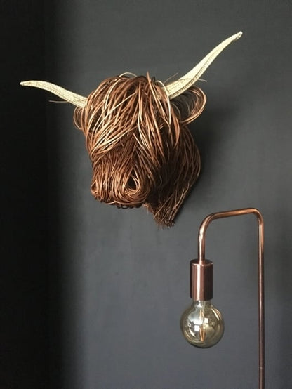 Geflochtener Kuhkopf mit hellen Hörnern an dunkler Wand neben einer Kupferlampe mit Glühbirne 1