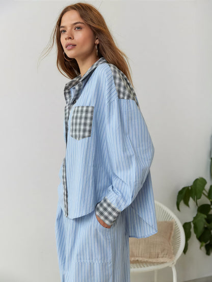 Model in blau gestreiftem Pyjama-Set mit grau kariertem Kragen und Brusttasche, seitlich vor weißem Korbstuhl und Zimmerpflanze. 3