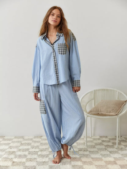 Ganzkörperbild im blau gestreiften Pyjama-Set mit weiter Hose, karierter Beintasche und Zugbändern an den Knöcheln, barfuß neben dem Korbstuhl. 7