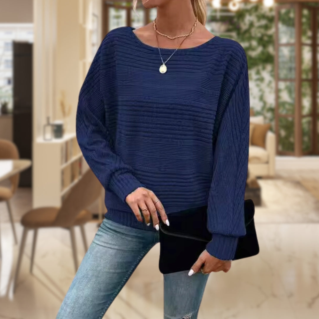 Frau in blauem Strickpullover und Jeans, hält schwarze Clutch, modernes Wohnzimmer im Hintergrund.