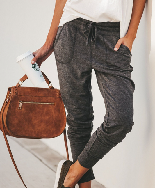 Frau in grauer Jogginghose mit brauner Ledertasche und Kaffeebecher, lässiger Streetstyle.
