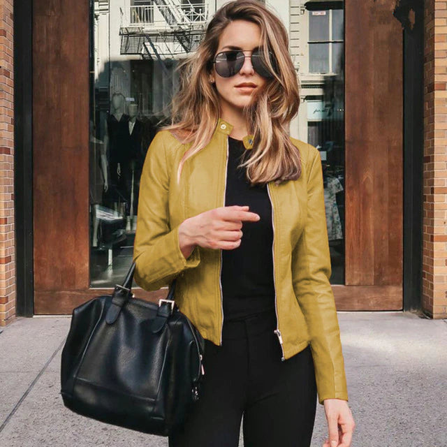 Frau in gelber PU-Lederjacke, schwarze Hose, Sonnenbrille, schwarze Tasche, urbaner Hintergrund.