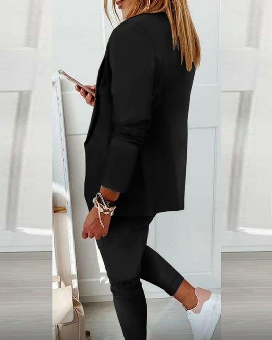 Frau in schwarzem Blazer und Hose, hält Smartphone, weiße Sneaker, moderne Damenmode.