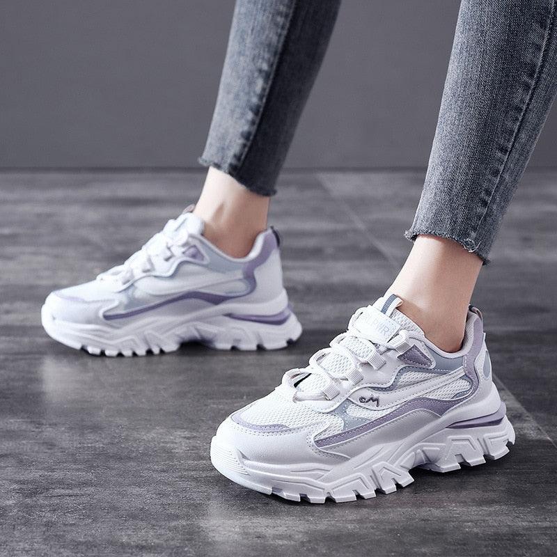 Weiße und lila Damen-Sneaker, klobiges Design, auf grauem Boden.