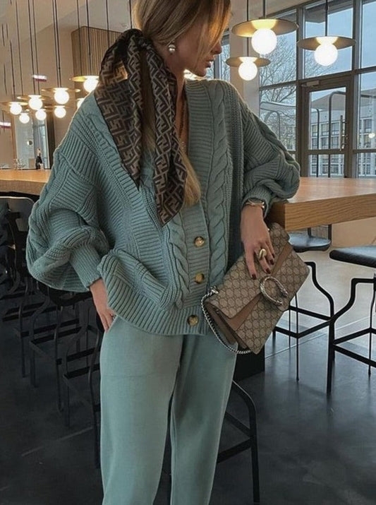 Frau in grünem Strickpullover und Hose, hält Designer-Handtasche, modernes Café-Interieur.