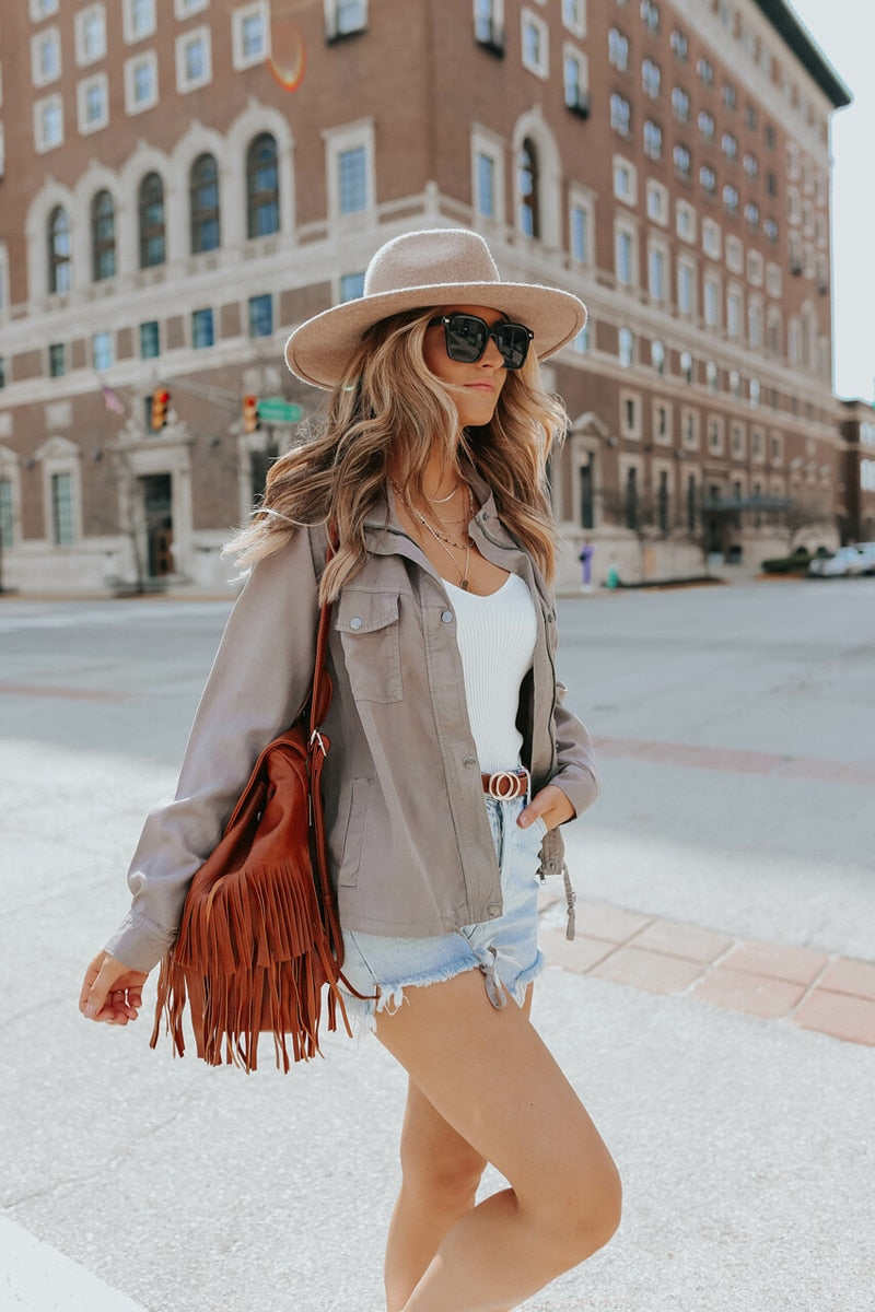 Frau in Sommeroutfit mit Hut, Sonnenbrille, Fransenledertasche, Jeansshorts auf Stadtstraße.