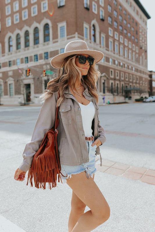 Frau in Sommeroutfit mit Hut, Sonnenbrille, Fransenledertasche, Jeansshorts auf Stadtstraße.