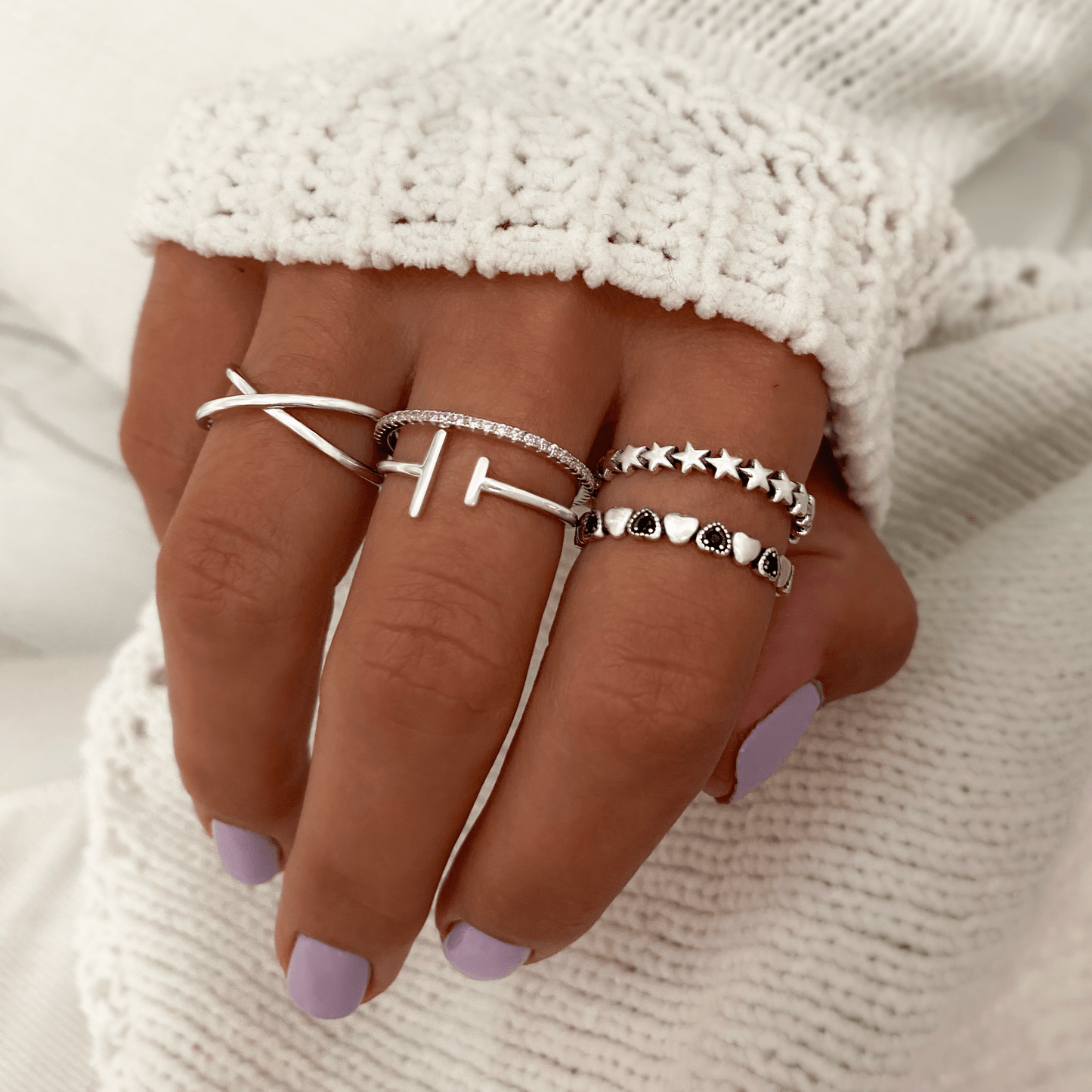 Hand mit vier silbernen Ringen, Kreuz- und Stern-Design, auf weißem Strickhintergrund.