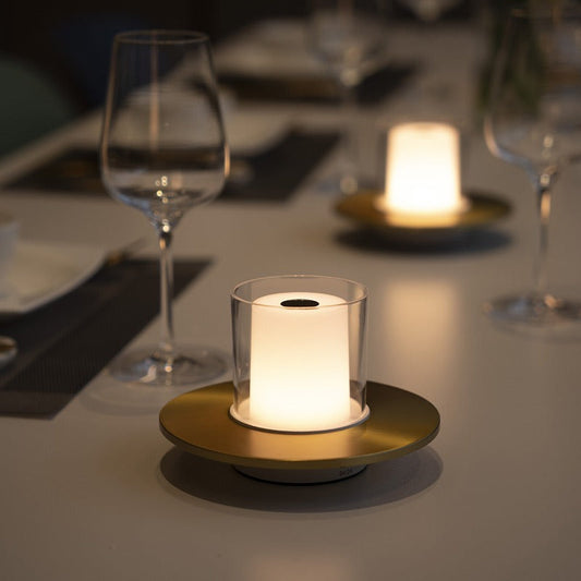 Moderne LED-Tischlampe mit Glaszylinder und goldener Basis auf elegant gedecktem Tisch.