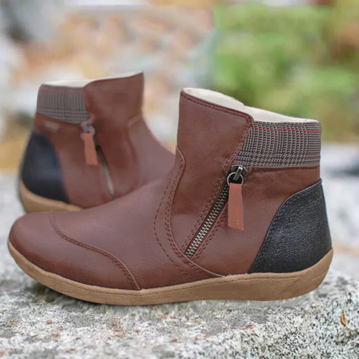 Braune Stiefeletten mit seitlichem Reißverschluss, kariertem Kragen und schwarzer Fersenpartie, draußen auf einem Stein vor unscharfem Hintergrund. 3