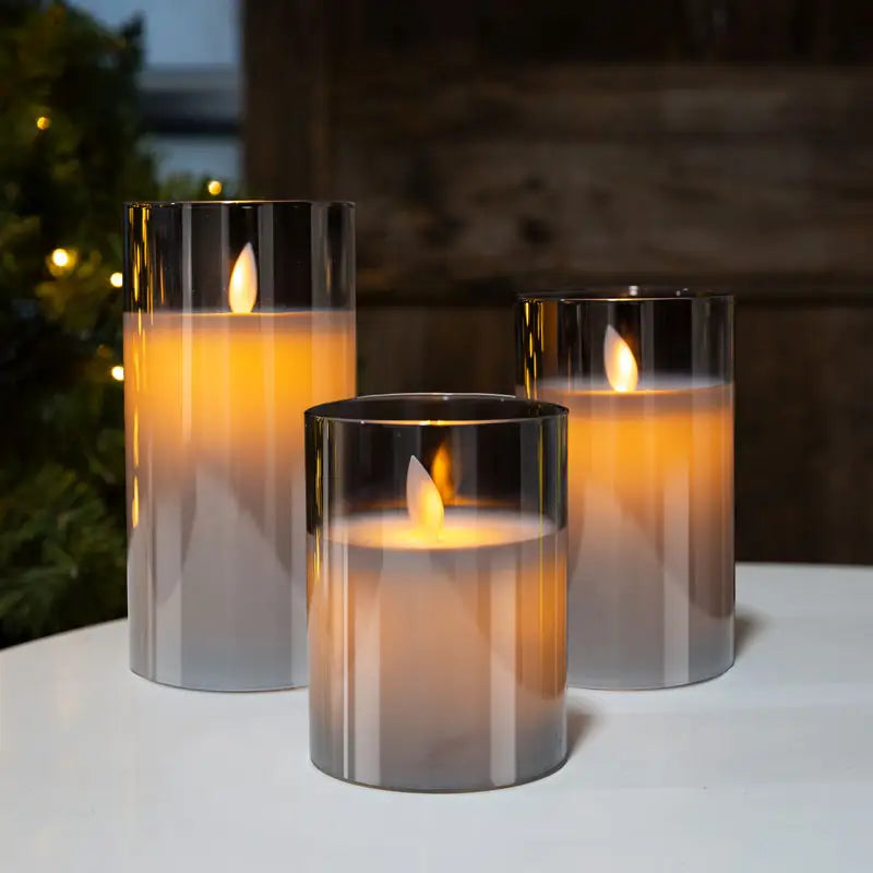 Drei LED-Kerzen in Glaszylindern, warmweiß leuchtend, auf weißem Tisch, dekorativ, modern.
