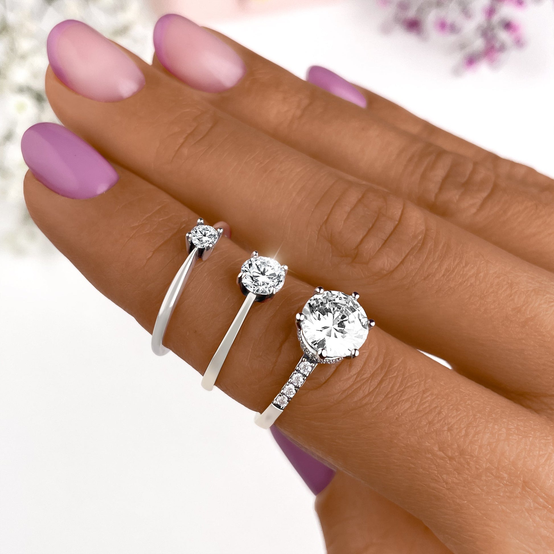Drei silberne Verlobungsringe mit Diamanten auf Hand, lila Nagellack, elegantes Schmuckdesign.