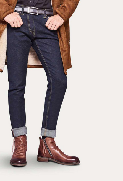 Mann in dunkelblauer Jeans, braunen Lederstiefeln und braunem Mantel, modisch gekleidet.