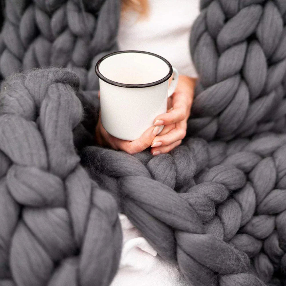 Frau mit grauer, grob gestrickter Decke hält weißen Emaillebecher. Gemütlich, warm, Winter.