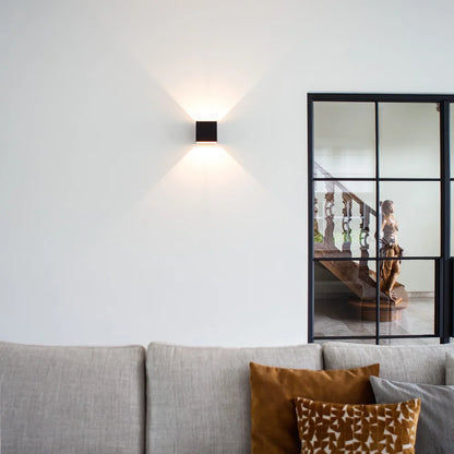 Schwarze Würfelleuchte an weißer Wand über einem grauen Sofa mit orangefarbenen Kissen, daneben eine Glastür mit schwarzem Rahmen. 10
