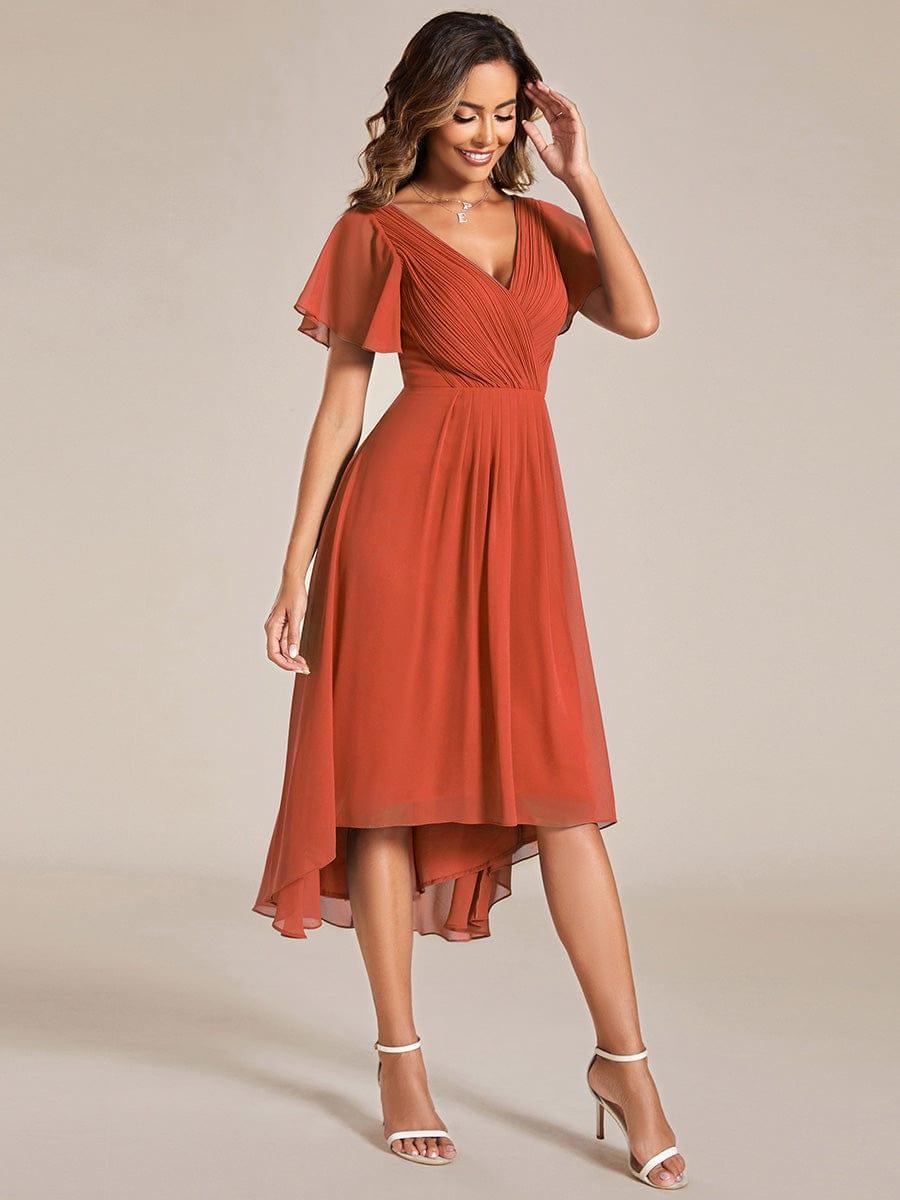 Orangerotes Chiffonkleid seitlich fotografiert, das Model fasst sich lächelnd ans Haar, der Rock schwingt locker mit hinterem Überlänge-Saum. 96