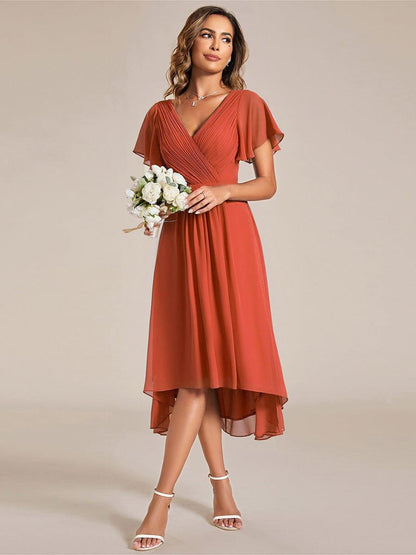 Model in orangerotem Chiffonkleid hält einen kleinen weißen Blumenstrauß, Blick zur Seite, vorne kürzerer Saum. 95