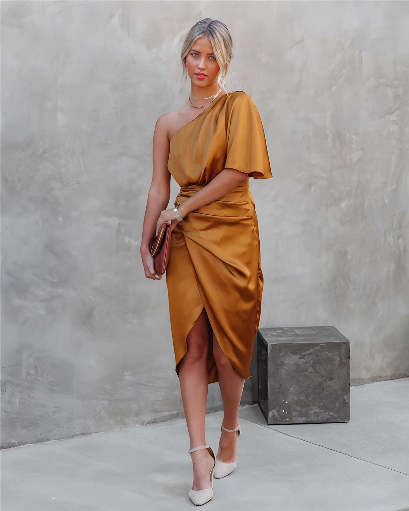 Frau in einem goldenen, schulterfreien Seidenkleid mit Clutch und weißen High Heels vor grauer Wand.