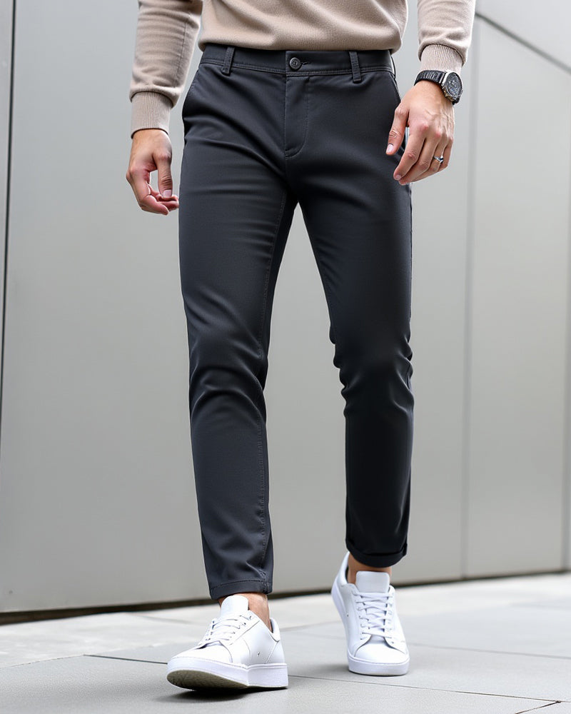 Anthrazitfarbene Chino mit beigem Strickpullover, Armbanduhr und weißen Sneakern vor grauer Wand, Beinsaum umgeschlagen. 9