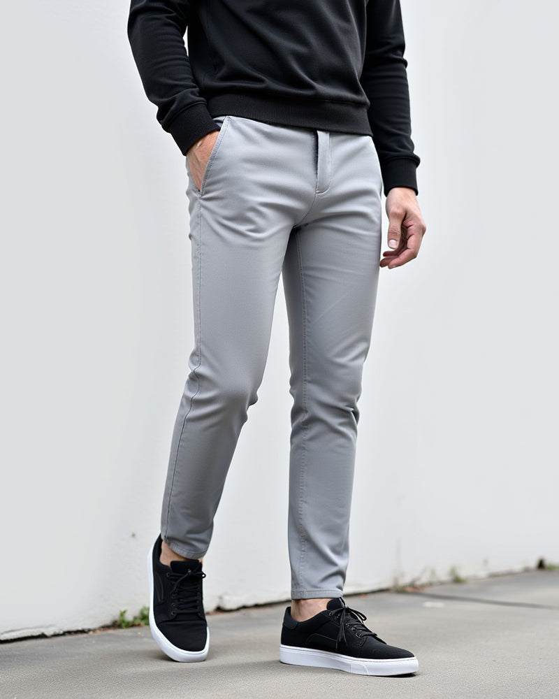 Hellgraue Chino im Freien vor einer weißen Wand, kombiniert mit schwarzem Pullover und schwarzen Sneakern mit weißer Sohle. 2